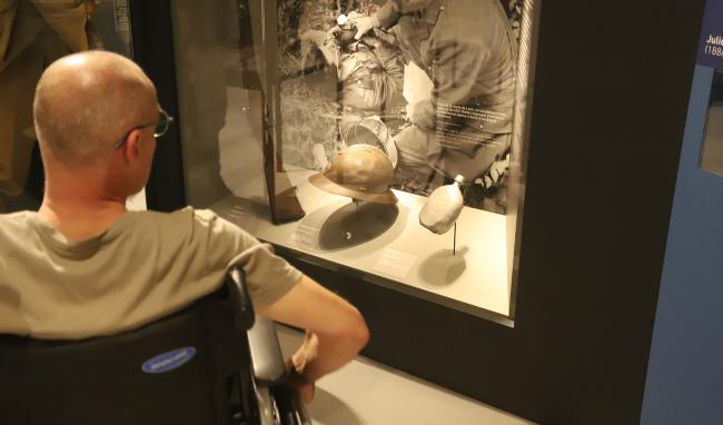 Personne en fauteuil roulant observant une vitrine présentant des objets historiques dans l’exposition du musée.